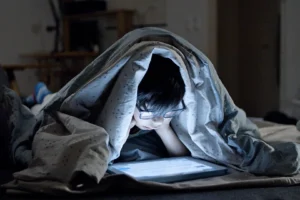 A young child wearing glasses lies under a blanket in a dark room, illuminated by the glow of a smartphone screen.